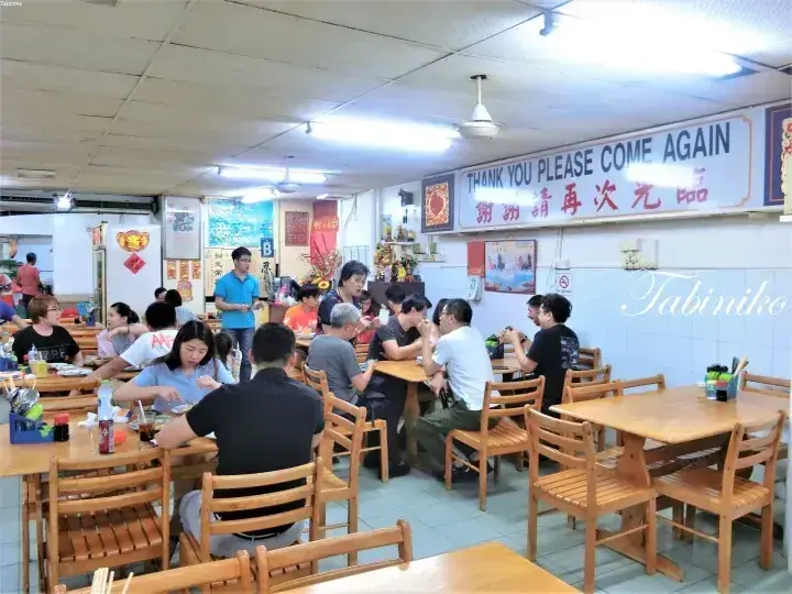 「THANK YOU PLEASE COME AGAIN」の看板が掲げられた賑やかな地元の食堂で、客が木製のテーブルで食事を楽しんでいる。