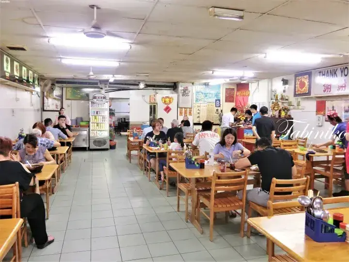 蛍光灯の下、木製のテーブルで食事をする客で賑わう伝統的なマレーシアのコピティアムの店内。