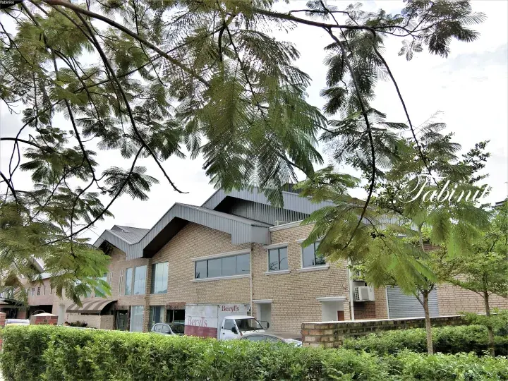 Modern brick building with angular roof and large windows, partially framed by lush green trees.