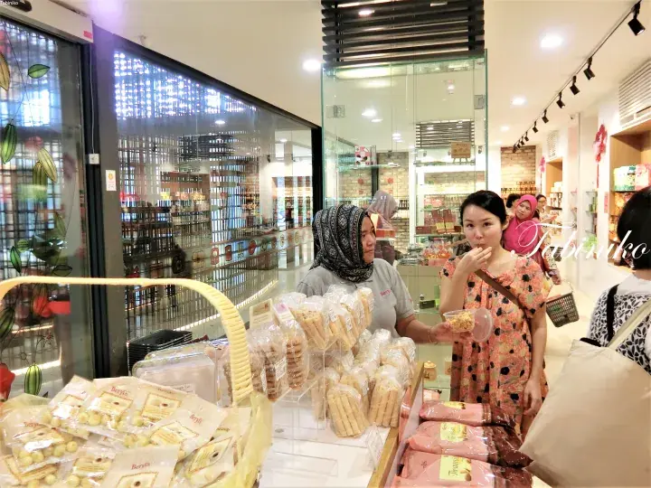 Customers sampling food at a display counter inside a modern Malaysian bakery or cafe.