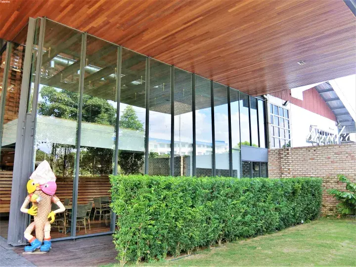 Modern glass-walled building with wooden ceiling and ice cream statue outside, surrounded by greenery.
