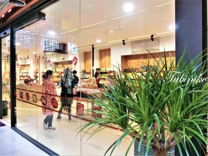 Shoppers browse inside a modern grocery store with glass storefronts and decorative plants outside.