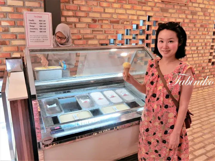 A woman holding an ice cream cone in front of a display case at a shop with a brick wall background.
