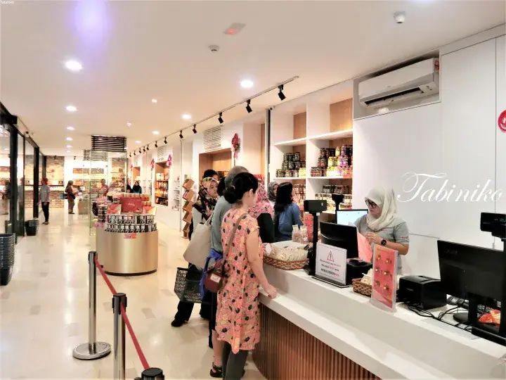 Customers queue at the checkout counter of a modern retail store with shelves of packaged goods and a staff member assisting.