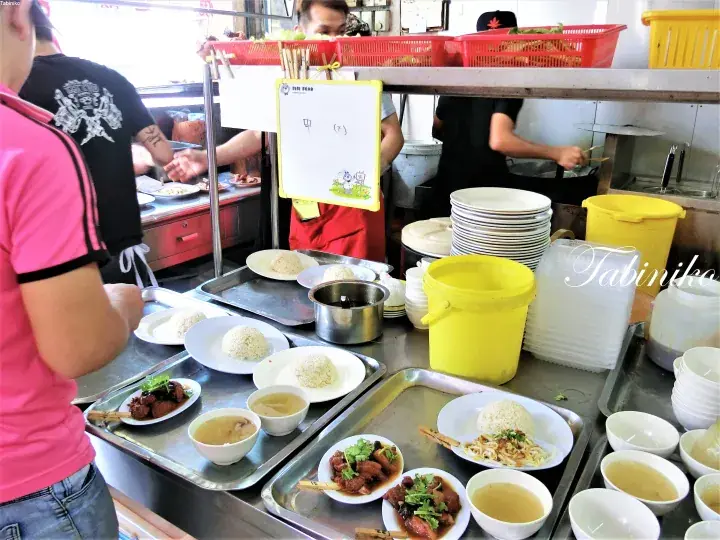 Busy food stall counter displaying plates of rice, meat dishes, and soup with yellow buckets and stacks of dishes.