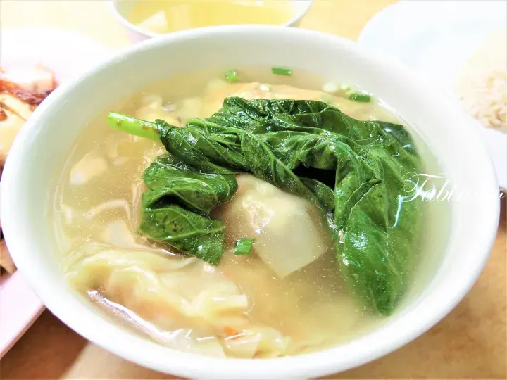 A bowl of clear wonton soup with fresh green vegetables and chopped scallions, a classic Malaysian comfort food.