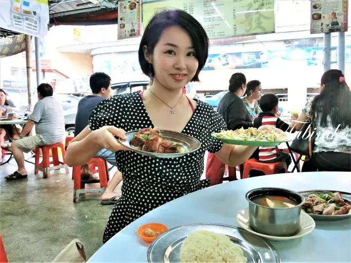 Woman holding plates of food at a bustling Malaysian hawker centre.