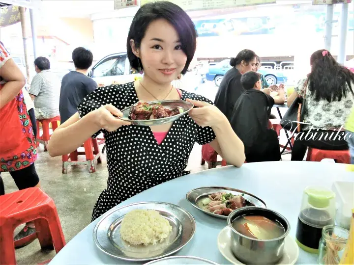 Smiling woman holding a plate of meat at a hawker centre table with rice and soup.