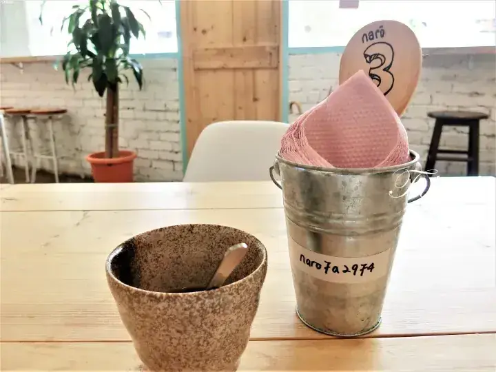 木製テーブルの上に置かれた陶器のカップと、ピンクのナプキンと「naro」の文字が書かれた木製サインが入った金属製のバケツ。