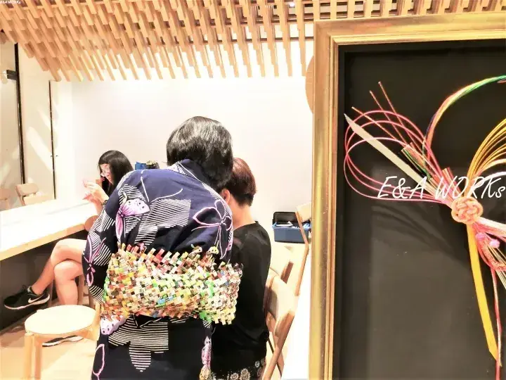 E&A WORKSで色鮮やかな帯を締めた人物と、右側のフレームに入った装飾的な蝶結びの展示。