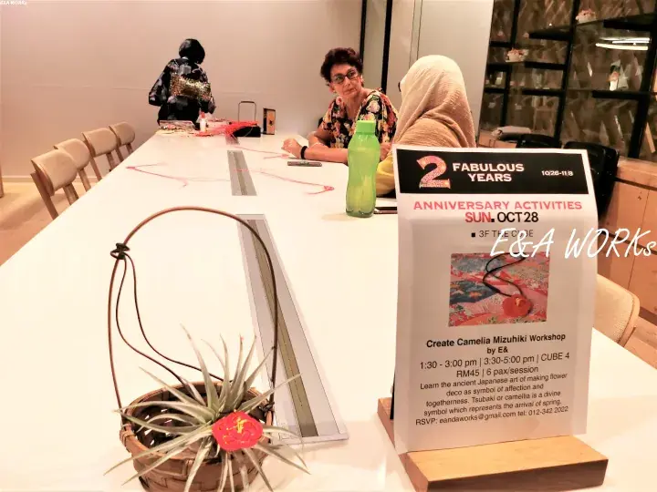 A workshop sign for Create Camelia Mizuhiki by E&I Works at a table with participants and a decorative air plant.