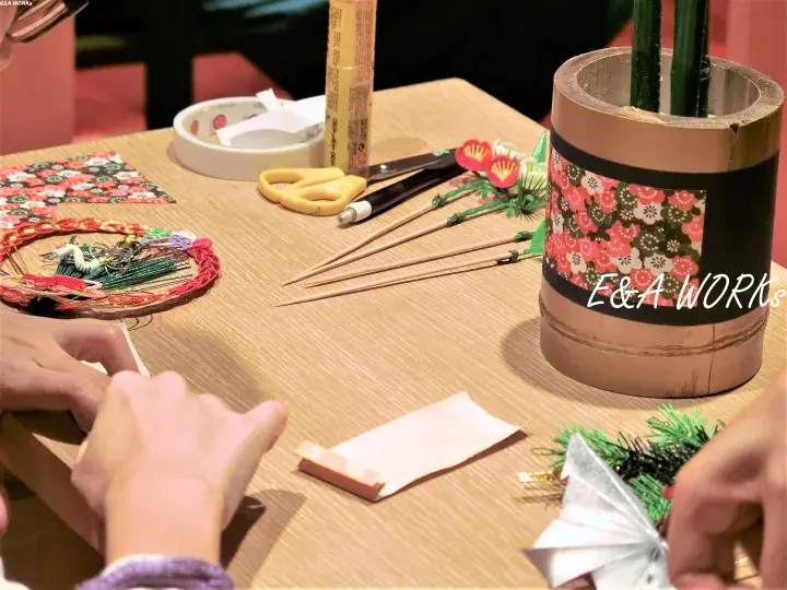 Hands crafting traditional Japanese decorations on a wooden table with bamboo, scissors, and floral paper.