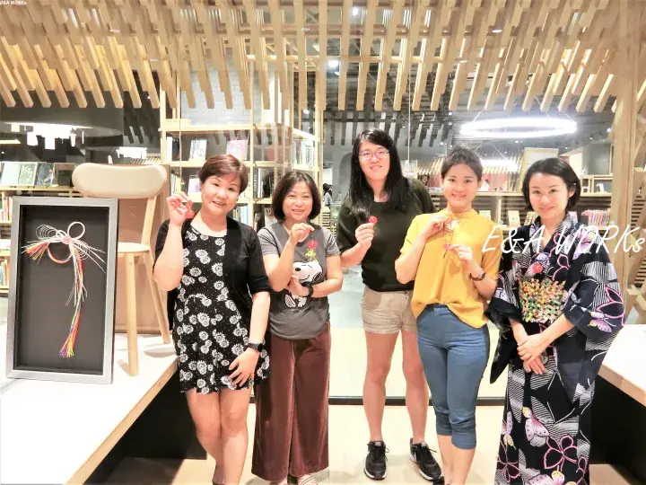 Five women posing with handmade crafts inside a modern retail space featuring wooden slat ceilings and display shelves.