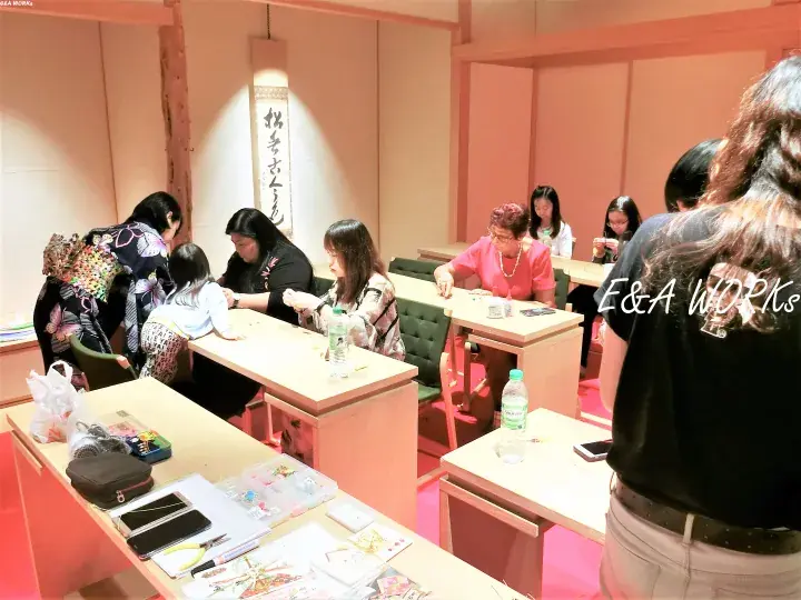 Attendees participate in a traditional craft workshop at E&A Works, seated at wooden desks in a warmly lit room.