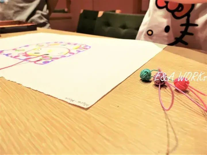 木製のテーブルに置かれたカラフルな絵と、手作りのゴムバンドのブレスレット。背景にはハローキティのシャツが見える。