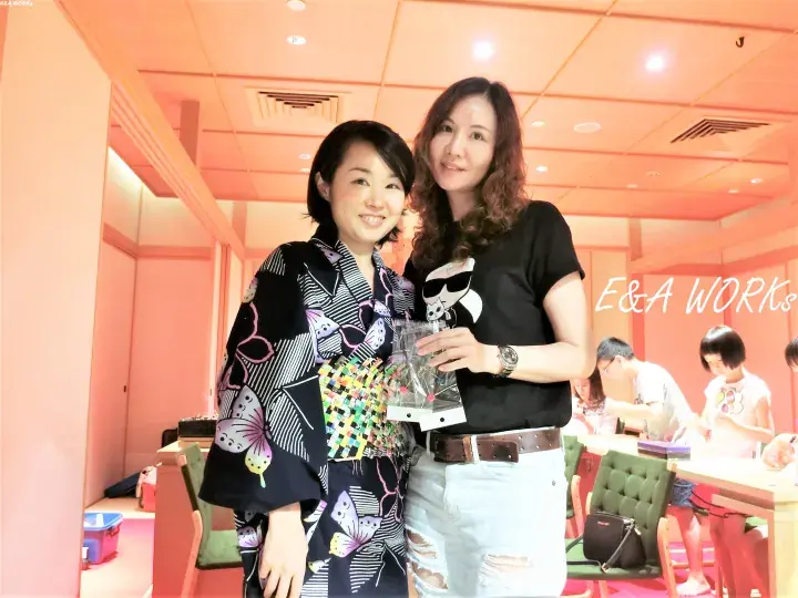Two women pose together indoors, one in a butterfly-patterned kimono and the other in a black t-shirt, at an event with E&A WORKS branding.
