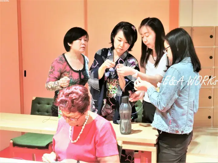A group of women participating in a craft workshop, focusing on threading materials together at a table.