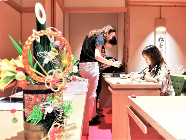 A woman writes at a wooden desk while another stands nearby, with a traditional Japanese flower arrangement in the foreground.