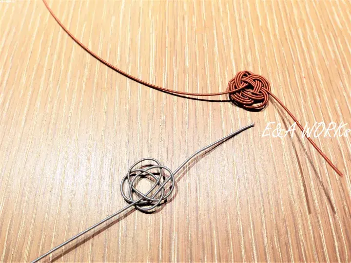 Close-up of two decorative knots made from copper wire and brown cord on a wooden surface, showcasing intricate craftsmanship.