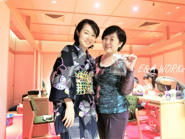 Two women smile at a workshop, one wearing a butterfly-patterned yukata and the other holding a small craft item.