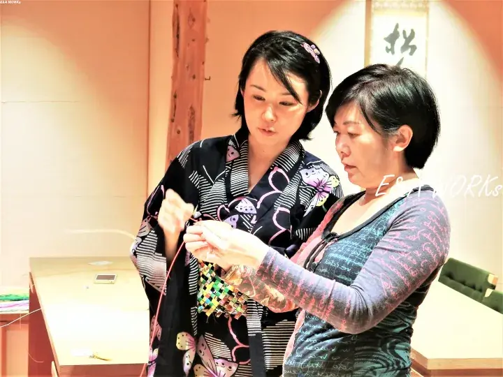 Two women collaborate on a traditional Japanese craft, with one wearing a patterned kimono and the other assisting with colorful threads.