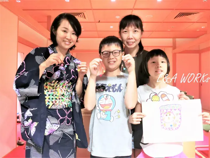 A family wearing handmade earrings poses in a pink room, with one child holding a drawing of a Doraemon-themed accessory.