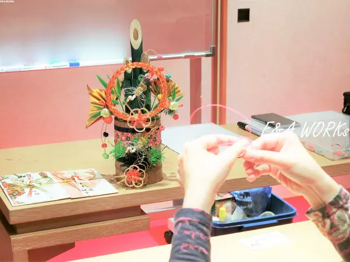 Hands crafting a traditional Japanese New Year decoration at a workshop, with a finished ornate arrangement displayed on a wooden table.