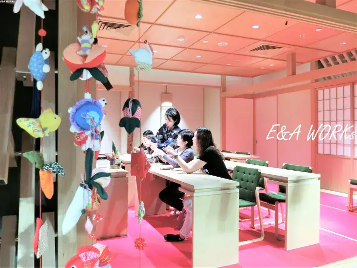 People crafting at a table in a pink room with hanging fabric decorations and E&A WORKs branding.