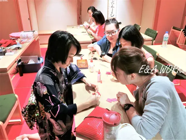 Women in a workshop crafting accessories at a table with pink decor and a sign reading さんくま