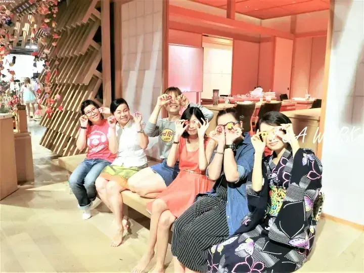 モダンな木製インテリアの店内ベンチで、友人たちが楽しそうにメガネを顔に当てている様子。