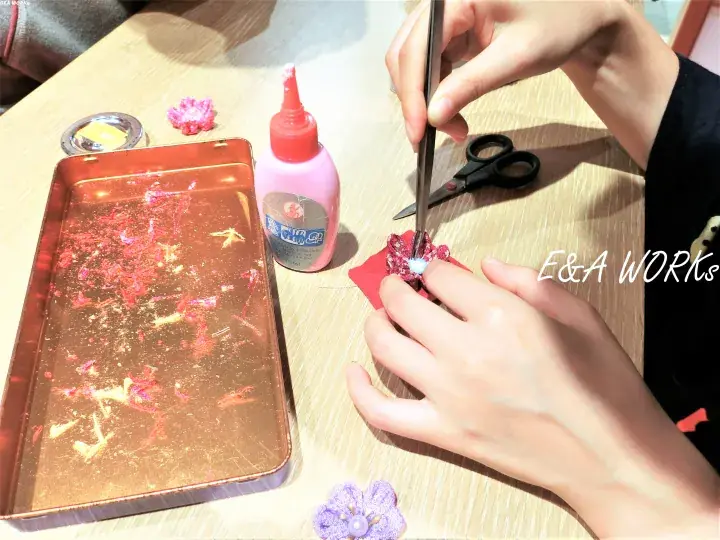 Hands using tweezers to assemble a flower craft with glue, scissors, and a tray of decorative materials on a wooden table.