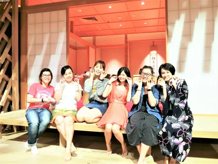 Six women posing playfully on a wooden bench inside a warmly lit Japanese-style restaurant interior.