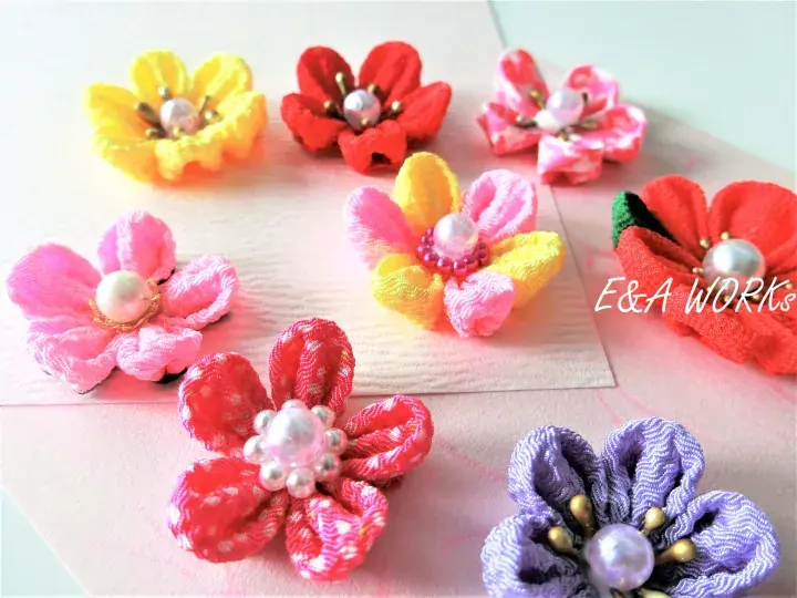 Colorful handmade fabric flowers with pearl centers arranged on a light pink surface, featuring the E&A WORKS watermark.
