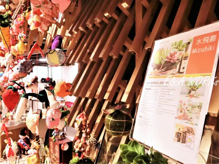 Colorful handmade ornaments hang near a wooden lattice wall beside a display board for Mizuhiki.