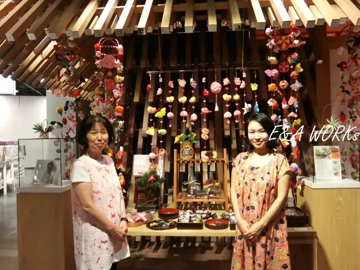 Two women stand beside a display of colorful hanging crafts and wooden decor at an E&A Works exhibition booth.