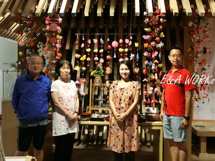 Four people posing in front of a wooden display featuring colorful hanging crafts and decorations.