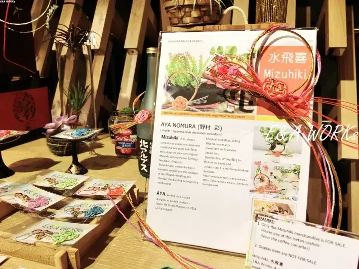 E&A WORKSのブースで展示された水飛喜の工芸品とAYA NOMURAのパンフレット、手作りアクセサリーが並ぶ様子。
