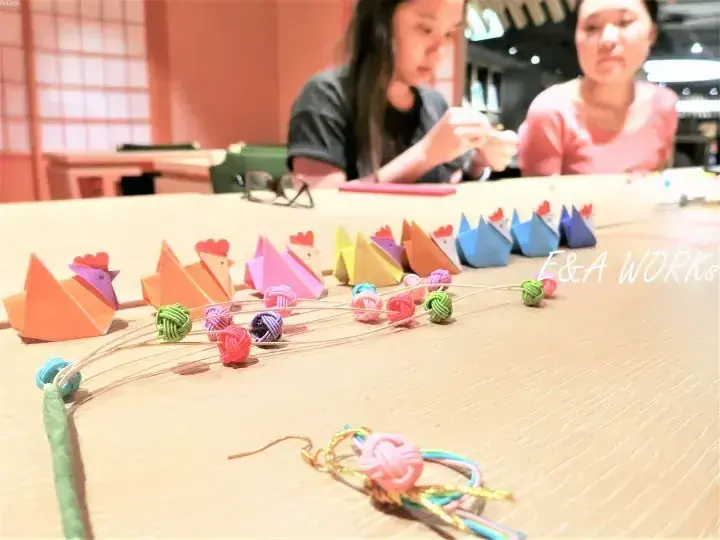 木桌上展示著色彩鮮豔的摺紙雞和編織工藝品,背景中有人在E&A WORKS工作。