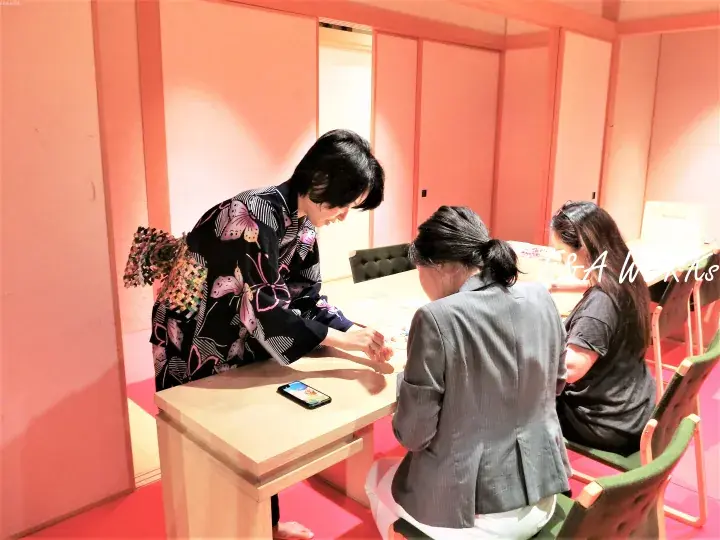 A woman in a black kimono with butterfly patterns writes at a table while two guests watch in a pink-walled room.