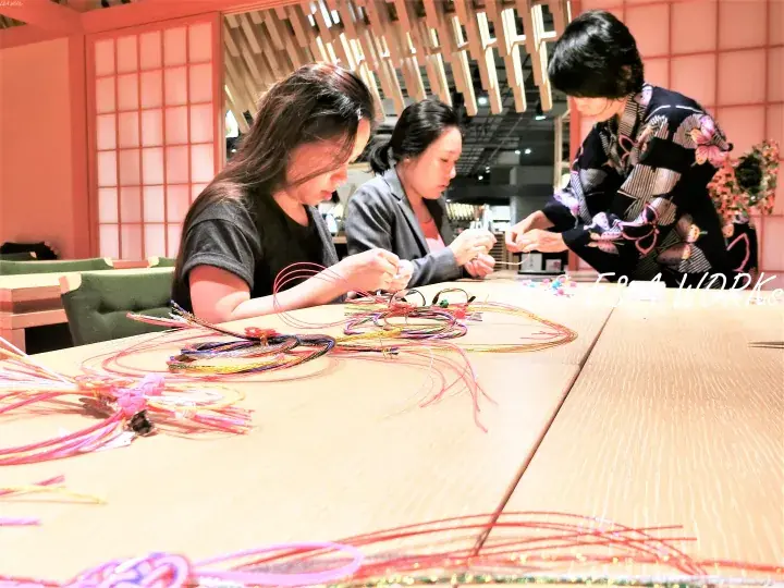 Three women crafting colorful woven bracelets at a wooden table in a modern Japanese-style interior.