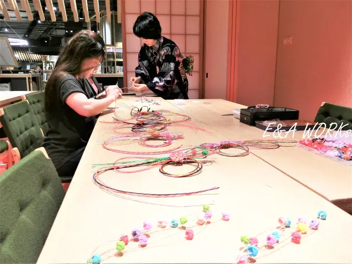 Two women crafting colorful bracelets at a long table in a modern workshop space with E&A WORKS branding.