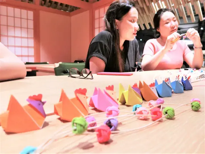 Two women crafting colorful origami rooster necklaces at a wooden table in a warm, modern interior space.