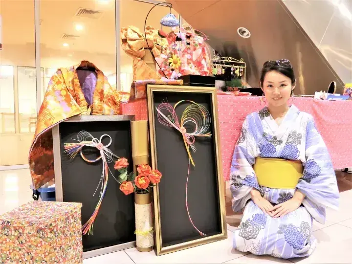 一位身穿藍白和服的女性跪坐在展示日本傳統工藝品和服飾的黑色展板旁。