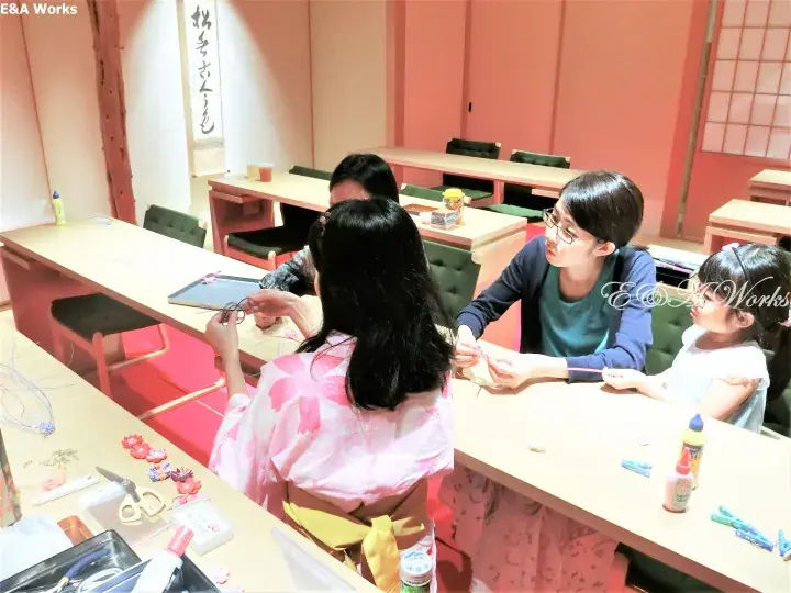 A woman in a pink kimono and a young girl participate in a traditional Japanese craft workshop with an instructor.