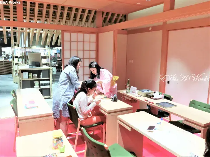 Three women interacting at a wooden desk inside a pink-themed pop-up space with shoji screens and bookshelves.