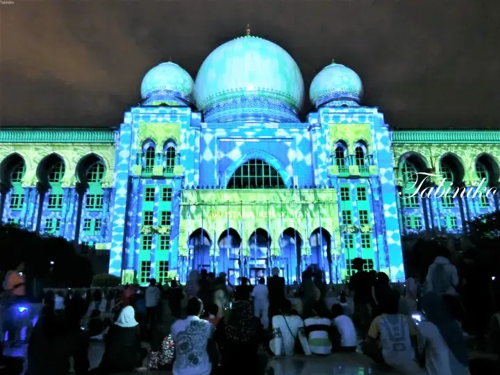 Putra Mosque illuminated with blue and green lights at night, surrounded by a crowd of visitors.