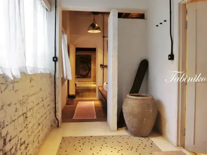 A rustic interior hallway with exposed brick walls, a large ceramic jar, and a view into a warmly lit bathroom with a pendant lamp.