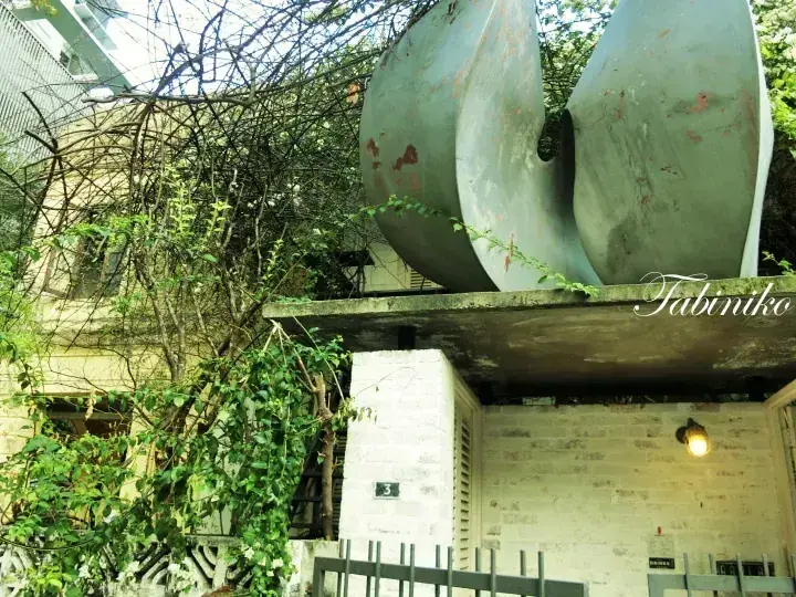 Large metallic sculpture resembling a propeller atop a concrete structure surrounded by lush greenery at a property entrance.