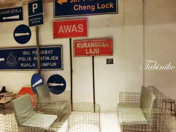Interior display of vintage Malaysian road signs including ‘AWAS’ and ‘KURANGKAN LAJU’ alongside wireframe furniture.