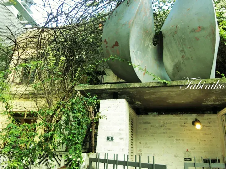 建物の入り口にあるコンクリートの構造物の上に設置された、緑豊かな植物に囲まれた大型の金属製プロペラ型彫刻。